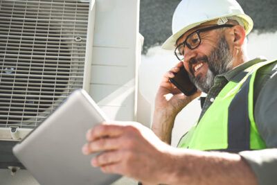 An HVAC business owner near a machine is reviewing fleet card usage data from his tablet while wearing a hard hat on the job.