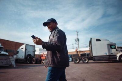 A happy truck driver outside his rig prepares to pay for fuel via his fleet card app.